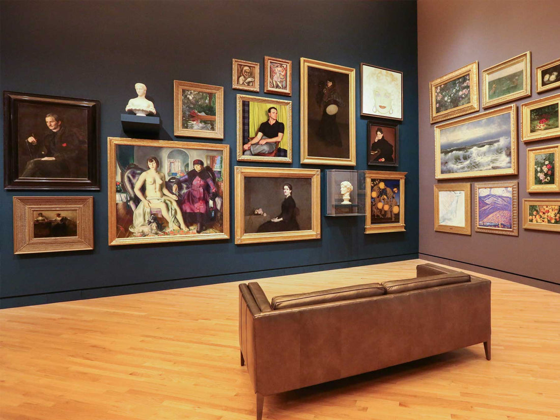 Historic paintings in ornate frames hung salon-style on a deep blue wall, with a bench